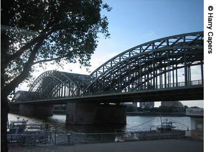 A side view of the Hohenzollernbrücke Bridge in Cologne, Germany. Two boats can be seen in the foreground in front of the bridge.