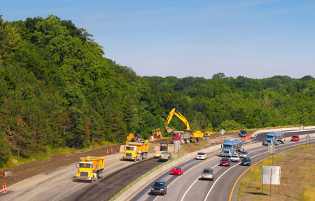 View of a highway with three lanes of traffic and an on-ramp in one direction; on the other side of concrete barriers there is construction activity with trucks, backhoes, loaders, a bulldozer, and a roller, working on additional lanes.