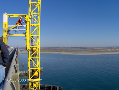 In the foreground, bridge inspectors are descending at the side of a bridge or causeway within a cage-enclosed ladder, part of equipment that sits on the bridge roadway and extends a caged-in platform up and horizontally over the barrier at the side of the bridge. Attached to the platform is a caged-in vertical ladder that extends well above and below the bridge surface. The bridge is quite long, disappearing into the horizon, spanning coastal waters.