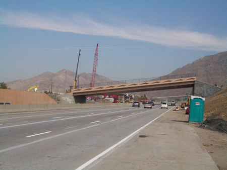 A view of the completed 4500 South Bridge in Salt Lake City, Utah. Construction cranes are visible in the background. Cars travel underneath the bridge.