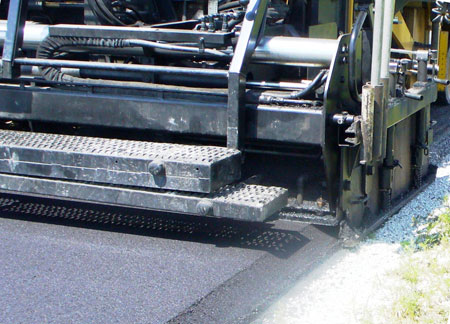 A close-up view of a paving machine implementing the Safety Edge on an asphalt pavement. The Safety Edge is a paving technique where the edge of the pavement is shaped to approximately 30 degrees.
