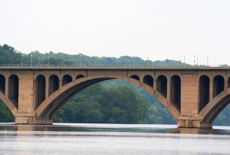 A side, ground-level view of an arch-type bridge spanning a river. A wooded area is in the background.