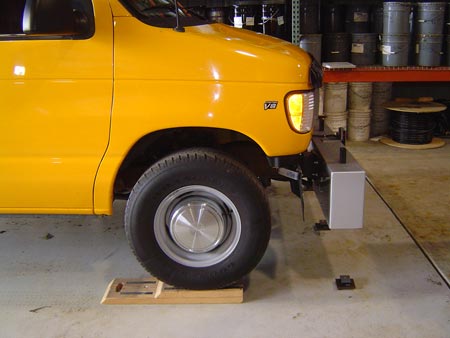 A closeup view of the height sensors of an inertial profiler being checked. A side view is shown of a van with the profiler attached in front. The van is positioned on wheel blocks, raising it up.