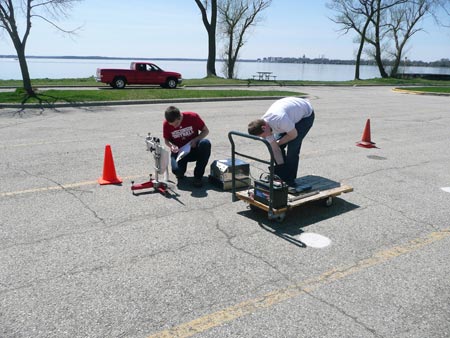 Two researchers at the University of Wisconsin-Madison's Modified Asphalt Research Center are preparing to use the Circular Texture Meter on a section of deteriorated asphalt pavement.