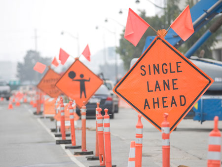 In the foreground is a traffic control sign that reads "Single Lane Ahead." In the background are other traffic control signs. Cars are driving away in the distance.