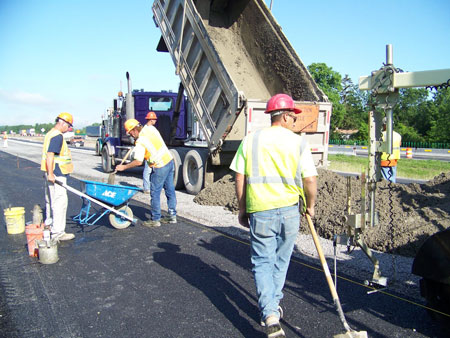 Three workers are in the foreground as long-life concrete pavement is constructed along a section of I-90 near Syracuse, NY. In the background is a dump truck that has just been unloaded.