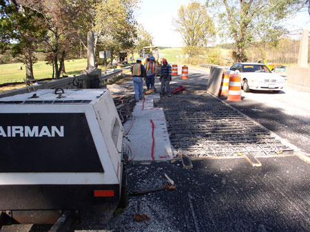 Workers repair a bridge deck. A bridge joint is visible. Three workers can be seen in the background. A car travels across the bridge in the next lane.