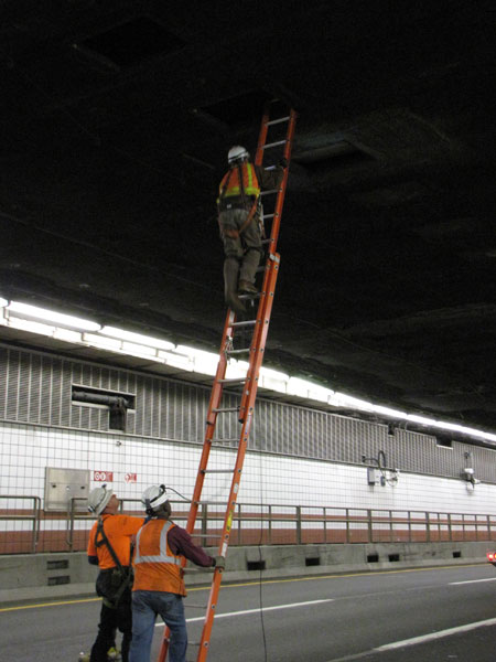 Three workers conduct a tunnel inspection. One worker is climbing a ladder to the roof of the tunnel. A second worker is holding the ladder, while a third worker stands next to him.