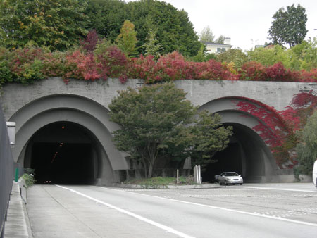 The entrance and exit portals of the Mount Baker Ridge Tunnel on I-90 in Seattle, WA. A car is exiting the tunnel. Trees grow on a median between the two portals.