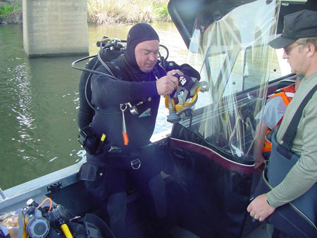 A diver in a wet suit with an oxygen tank strapped to the back of the suit is on a boat, preparing to go under water to inspect a bridge. The diver is holding a camera. To the right is another member of the inspection team on the boat.