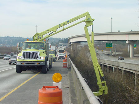 A truck parked on the side of a highway bridge lowers under-bridge inspection equipment. Traffic is traveling to the right of the truck. Orange construction barrels line the side of the bridge.
