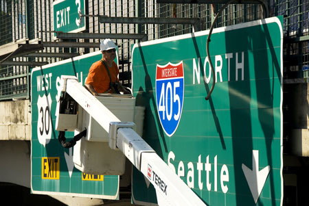 A worker is in a mechanical lift in front of an overhead highway sign. The sign reads: Interstate 405 North Seattle.