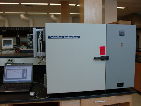 A view of the Asphalt Binder Cracking Device in a laboratory. The device is on a counter with a computer next to it. In the background are other computers.
