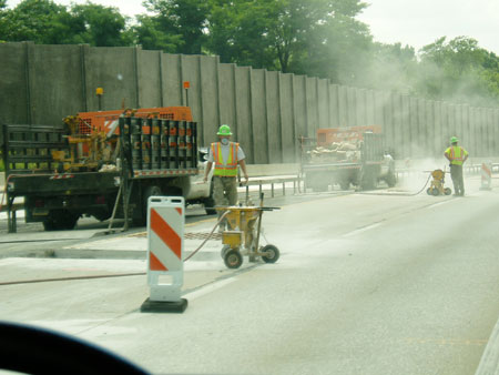 Two workers wearing safety vests, hard hats, and face masks in a work zone. Two trucks are in the background. Dust is rising up from the pavement.