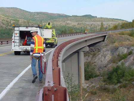 A bridge inspector wearing a safety vest and hard hat walks along the shoulder of a steel girder bridge. Two trucks with equipment are in the background.