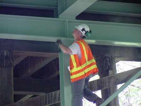 An inspector wearing a safety vest and hard hat climbs up a steel beam to inspect a bridge's steel substructure unit.