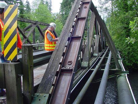 An inspector wearing a safety vest and hard hat looks down as he visually examines a truss structure.