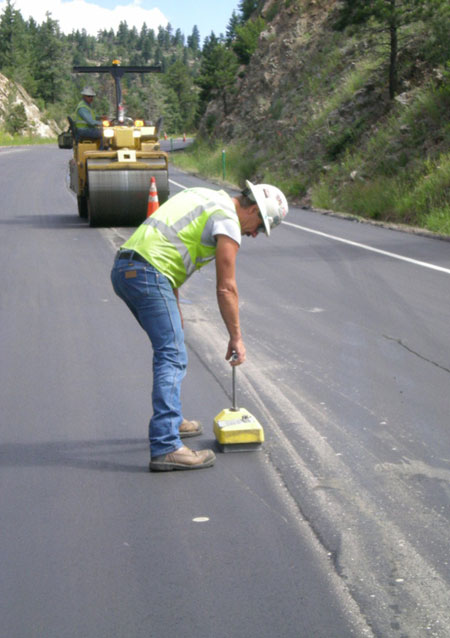 A highway worker examines an asphalt pavement joint during paving. In the background is a roller with a driver sitting on it.