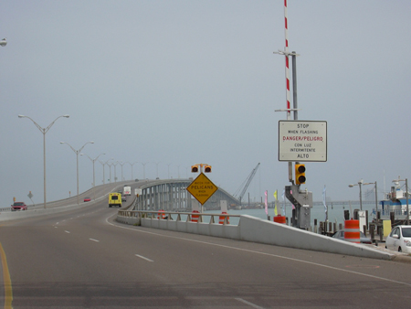 A view of the approach to the Queen Isabella Causeway bridge in Texas. To the right in the foreground is a sign mounted to a traffic gate. The sign reads "STOP WHEN FLASHING. DANGER/PELIGRO." Traffic is crossing the bridge in both directions.