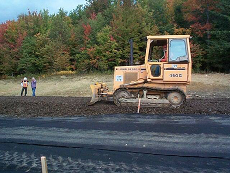 A road worker operating a bulldozer places tire-derived aggregate on a roadway. Two additional workers are in the background.