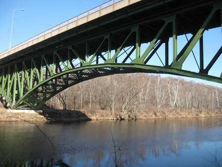 A ground-level view looking up from the under-side of a steel deck truss bridge in Sandstone, MN. The bridge carries State Road 123 over the Kettle River. A wooded area is in the background.