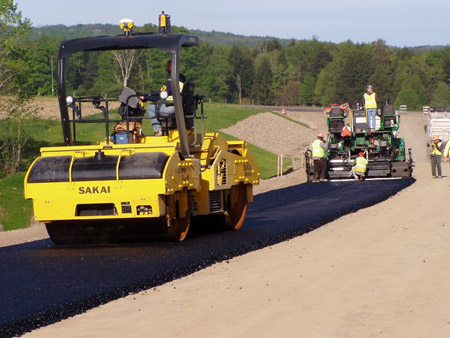 A view of an intelligent compaction roller placing a hot-mix asphalt lift on U.S. 219 in Springville, NY. Behind the roller is other paving equipment, with workers riding on the equipment and standing in front of it.