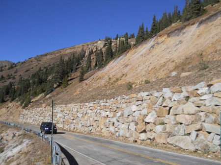 A car travels along a two-lane roadway next to a rockery wall at the toe of a scaled rock and soil cut slope along Guanella Pass Road near Georgetown, CO. In the background is a hilly area with trees.