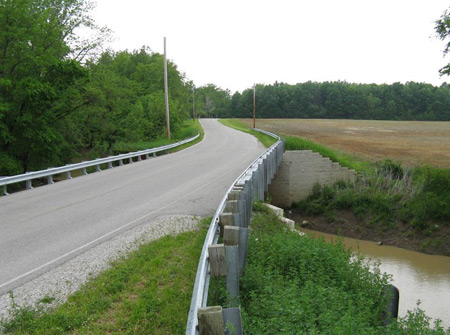 View of a two-lane bridge with guardrail over a narrow creek in a rural area. Farmland is on one side and a wooded area on the other.