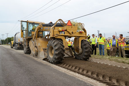 Highway engineers observe a demonstration of in-place recycling equipment on a roadway.