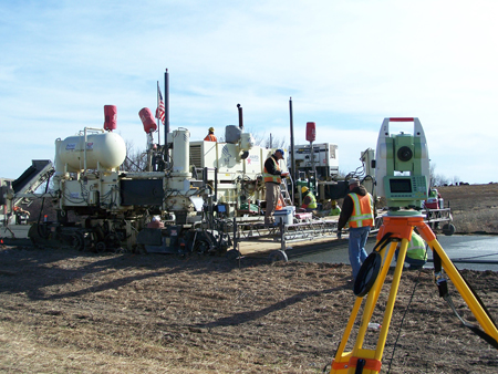 A view of a stringless concrete paving construction site. Paving equipment and six highway workers wearing orange and yellow safety vests are visible.