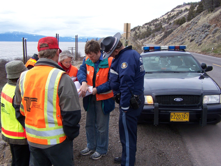 A road safety audit team in Oregon stands on the side of a mountainous roadway, next to a police car. The six members of the team are looking at documents.