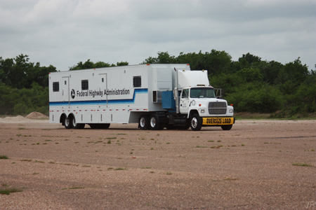 A view of the outside of FHWA's Mobile Asphalt Testing Lab. The lab is housed in a tractor-trailer truck with the FHWA logo and "Federal Highway Administration" displayed on the side of the truck. On the front bumper of the truck is a banner that reads "Oversize Load."