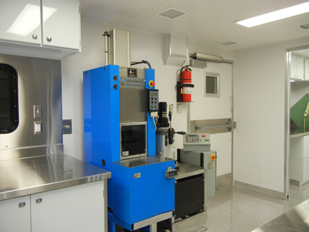 An interior view of FHWA's Mobile Asphalt Testing Lab. In the foreground is a gyratory compactor. To the left of the gyratory compactor is a stainless steel counter. To the right is a fire extinguisher mounted on the wall.
