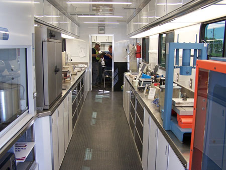 A view of the interior of FHWA's Mobile Concrete Laboratory. The lab is lined on each side with stainless steel counters holding pavement testing equipment. Underneath the counters are cabinets and drawers. On the right-hand wall in the background is a red fire extinguisher. Two lab personnel are also visible in the background.