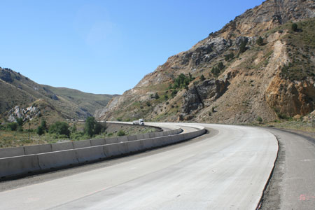 A highway built with concrete pavement in a mountainous area. A truck and cars are seen in the background about to round a curve in the roadway. To the right above the highway is a rocky slope.