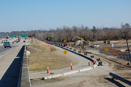 A view of the Neches River Bridge Project in Texas. To the left, traffic is traveling on a highway. To the right is a construction area with four orange barrels in the forefront and additional barrels in the background, along with sections of pipe.