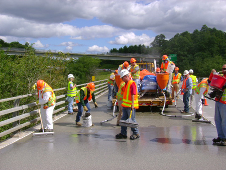 A bridge maintenance crew seals and waterproofs a reinforced concrete bridge deck. Twelve workers wearing safety vests and hard hats are visible. Workers are using mops to apply sealants and overlays. Three workers in the background are on a flatbed truck, controlling the flow of materials to the deck through a hose. In the farther background is a highway overpass. To the right is a wooded area.