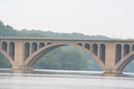 Partial side view of a large, masonry, open-spandrel, multiple-arch bridge over still water with guardrail and streetlights atop bridge. A forested hillside is in the background.