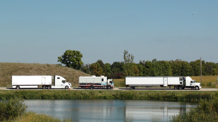 One truck and two tractor-trailers containing moving pavement deflection devices are seen traveling on a roadway next to a pond. A wooded area is in the background.