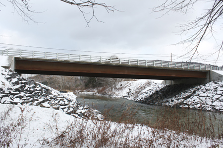 The Route 23 bridge in Oneonta, NY, as seen from the side of the bridge in winter time. Snow is on the banks of the waterway running under the bridge. The single-span bridge was built using precast concrete deck panels and field-cast ultra-high performance concrete connections.