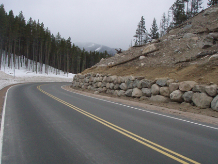 Bear Lake Road in Colorado's Rocky Mountain National Park in winter. Snow is on the ground. To the right of the roadway is a slope with a rockery at the bottom of the slope containing stacked angular rocks. To the left in the distance is a wooded area.