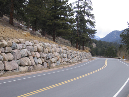 A view of Bear Lake Road in Colorado's Rocky Mountain National Park. To the left of the roadway is a slope with a rockery at the bottom of the slope next to the roadway containing stacked angular rocks. Behind the rockery and in the distance are wooded areas.