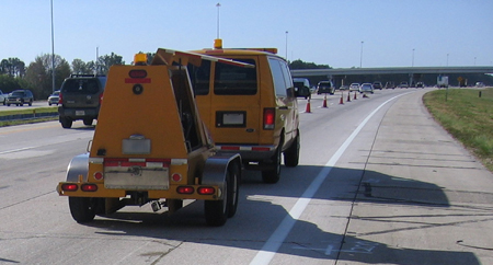 A rear view of a falling weight deflectometer (FWD) being towed by a van on a highway. A row of traffic cones separates the FWD from the other two lanes on the highway, which are to the left of the lane the FWD is traveling in. Traffic is moving in the other two lanes. Up ahead is an underpass.