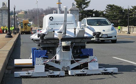 The image shows the RABIT™ bridge deck assessment tool on the Arlington Memorial Bridge. A car is shown passing to the right of the RABIT™ bridge deck assessment tool, and a crane-type truck is shown behind the RABIT™ bridge deck assessment tool.