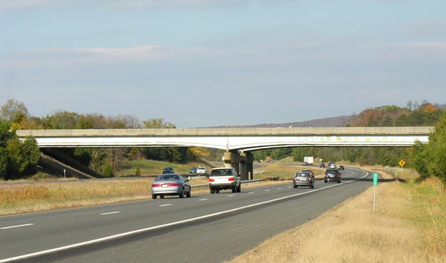 This photo displays a view of the Virginia Pilot Bridge in Haymarket, Virginia as shown from Interstate 66. The bridge spans horizontally across the image, and cars are passing underneath the bridge in both directions while travelling on a highway.