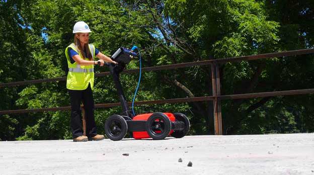 Figure 2. Photo. Data collection on a bridge deck using ground-coupled GPR. In this figure, a worker is pushing a ground penetrating radar unit on the deck of a bridge. The unit has two wheels in front and two in the rear. A red plastic box between the wheels is in contact with the deck surface. Above the red box is a rectangular control unit resting at an angle on the framework extending from the wheels to the worker’s hands. A computer is at the top of the unit, almost at the worker’s eye level.