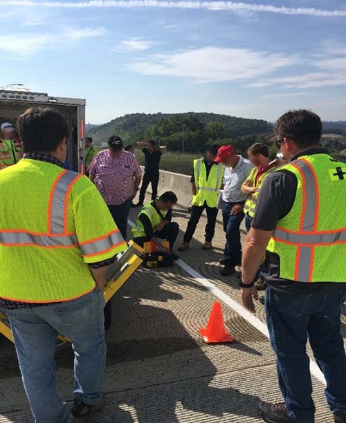 This photo shows approximately ten people on a concrete bridge surface. Seven people are wearing safety vests. The group is gathered around a man kneeling on the surface, demonstrating the evaluation of a bridge deck.