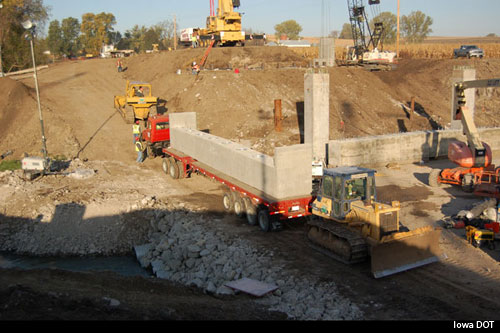 This photo shows the construction site of a bridge being built using PBES components. The bridge is on U.S. Route 6, traversing Keg Creek in Iowaâ€™s Pottawattamie County. A truck is carrying a prefabricated abutment with corrugated voids (or pile pockets), which are backfilled with self-consolidating concrete after the abutment is set.