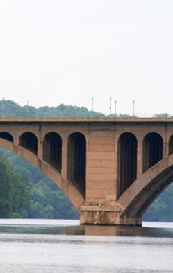 Figure 2. Photo. A side, ground-level view of an arch-type bridge spanning a river. A wooded area is in the background.