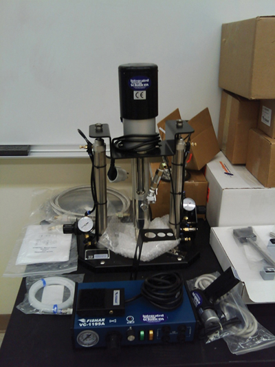 A new SprayWriter is shown on a table surrounded by various components (manual, plastic tubing, metallic hose with brass fitting, other attachments) wrapped in plastic. The equipment appears to be about 3 ft high on a base of approximately 4 to 6 sq ft. Part of the unit is labeled FISNAR VC-1195A. Visible parts of the equipment include three dials, switches, a hose, a power cord, and others. Some structural components appear to be made of stainless steel.